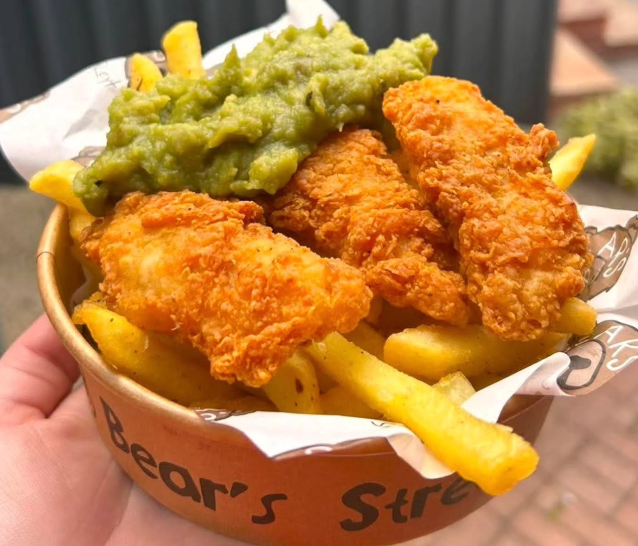 A round cardboard container filled with golden fried fish goujons, thick chips and mushy peas for TV & film catering.