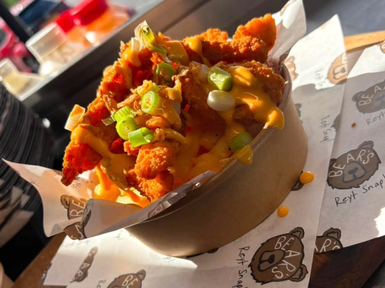 A cardboard bowl filled with golden fries topped with crispy chicken tenders, melted cheese sauce and spring onions for TV & film catering.