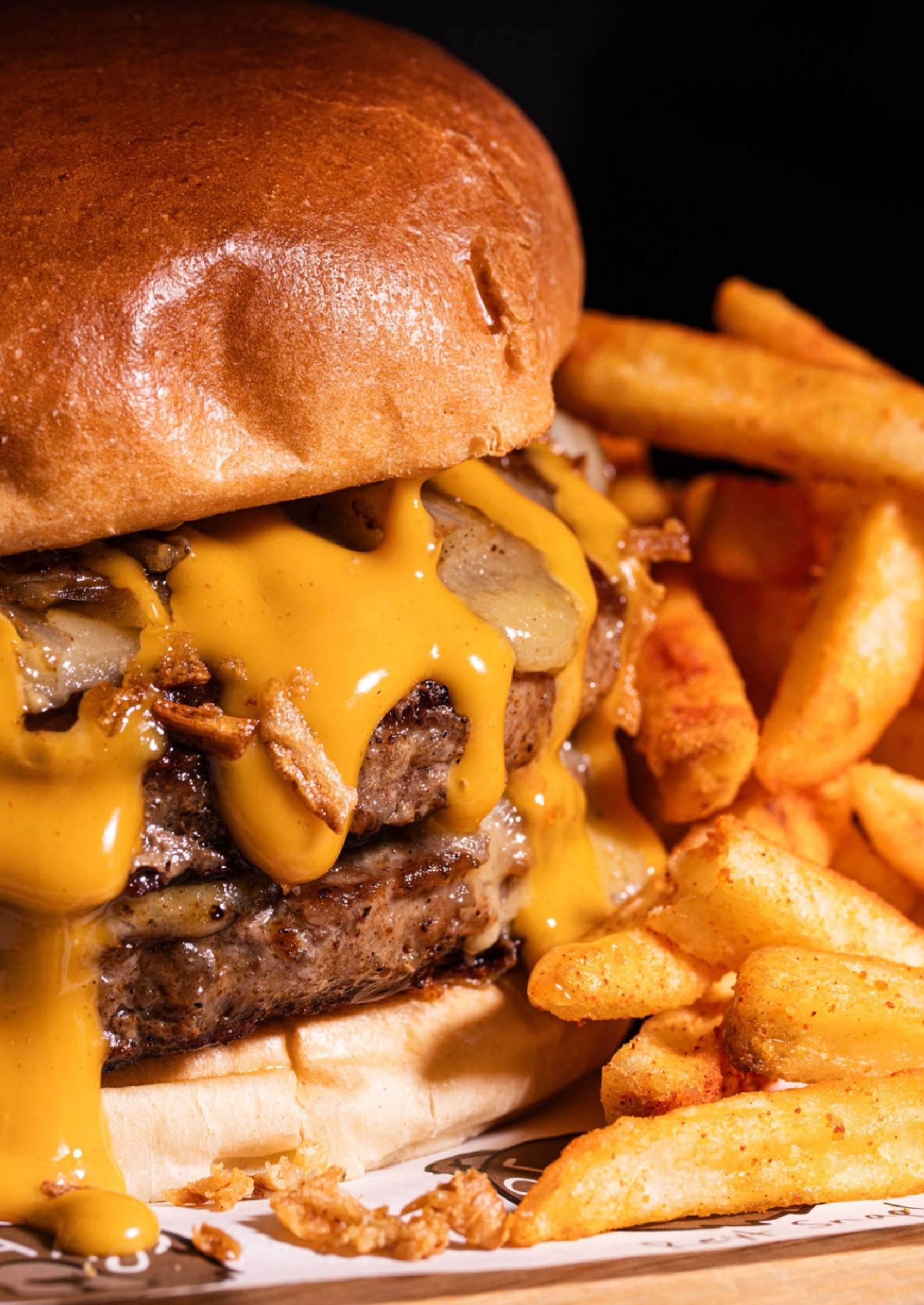 A massive multi-layered live-fire smash burger with melted cheese and onions on a wooden serving board, representing premium hot menu upgrades available through workplace canteen catering.